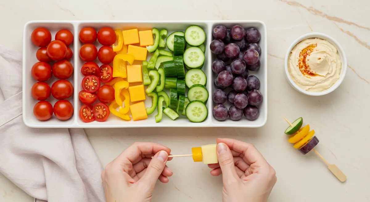 Vibrant rainbow sandwich skewers with cheese, veggies, and hummus dip, arranged in color order on a light background