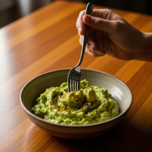 A hand holding a fork over a bowl of mashed avocado for making guacamole.