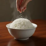 How to Cook Perfect Stove Rice in 20 Minutes: Foolproof Method for Easy, Fluffy Results A hand sprinkles salt over a steaming ceramic bowl of fluffy white rice on a dark wood table.