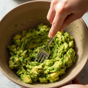 Mashing avocado with lime juice and salt in a ceramic bowl using a fork.