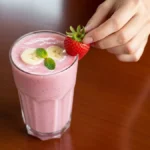 A glass of the best strawberry banana smoothie for kids, with a hand placing a strawberry on the rim.