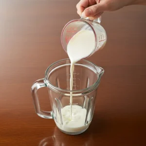 A hand pouring milk into the bottom of a blender jar.