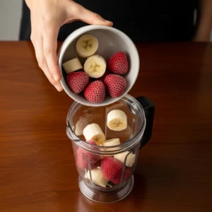 A hand adding frozen strawberries and bananas to a blender.