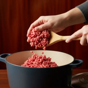 A hand crumbling and browning ground beef in the Dutch oven.