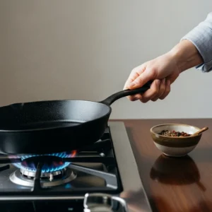 A hand holding a cast-iron skillet over a gas flame to heat it up for cooking.