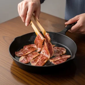 A hand using tongs to place raw, marinated beef slices into a hot, empty cast-iron skillet to begin cooking.