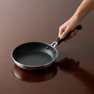 A hand holding the handle of a non-stick frying pan on a dark wood table.