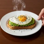 A perfect avocado toast with a sunny-side-up egg, garnished with everything bagel seasoning and microgreens, on a dark wood table.