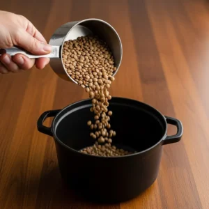 A hand pouring a cascade of rinsed brown lentils from a measuring cup into a pot.