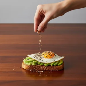A hand sprinkling everything bagel seasoning over the finished avocado toast with egg.