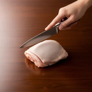 A chefs knife held over a boneless chicken thigh on a cutting board.