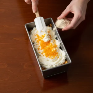 A hand using a spatula to fold fresh orange zest into the creamy cottage cheese layer in a loaf pan.