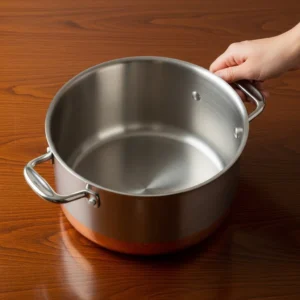 Beautiful large heavy-bottomed stainless steel pot with copper core sitting empty on dark wood table with human hand on handle.