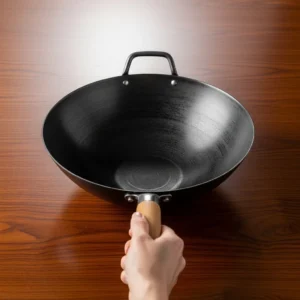 Dark carbon steel wok with a human hand gently holding the handle on a dark wood table