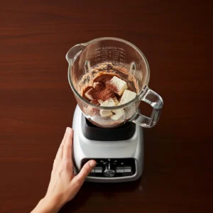 High quality glass blender on dark wood table containing chocolate mousse mixture with tofu and cacao visible inside, human hand resting on base
