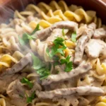 Close-up overhead view of a rustic ceramic bowl filled with creamy beef stroganoff, showing tender beef strips and mushrooms coated in thick sauce, garnished with fresh parsley, with light steam rising.