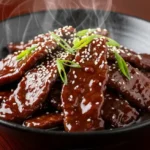 Close-up of glossy Mongolian beef in a dark ceramic bowl with steam rising and sesame seeds scattered on top.