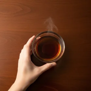 Small glass bowl of maple syrup on dark wood table, human hand holding bowl from underneath, tiny wisp of warmth rising subtly visible