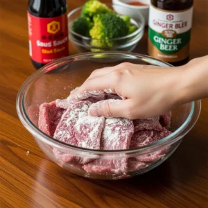 A human hand massaging cornstarch into sliced beef in a glass bowl for the velveting technique.