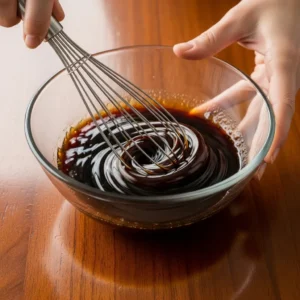 Human hand whisking dark brown sugar and soy sauce in a glass bowl creating glossy swirls