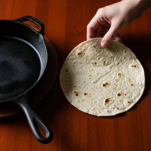 A warm corn tortilla is being placed on a dark wood table next to a skillet for serving breakfast tacos