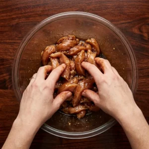 raw chicken strips marinating in coconut aminos inside glass bowl with feminine hand mixing